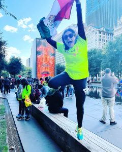 Teresita saltando y alzando la bandera de México. Porta una pants negro y una playera verde que dice: Achilles México